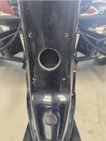 Close-up of a black motorcycle frame with a large circular cutout and several small holes for mounting a component.