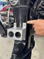 Metal mounting block attached to a motorcycle frame with two circular holes; a person's finger points to the gap beside it.